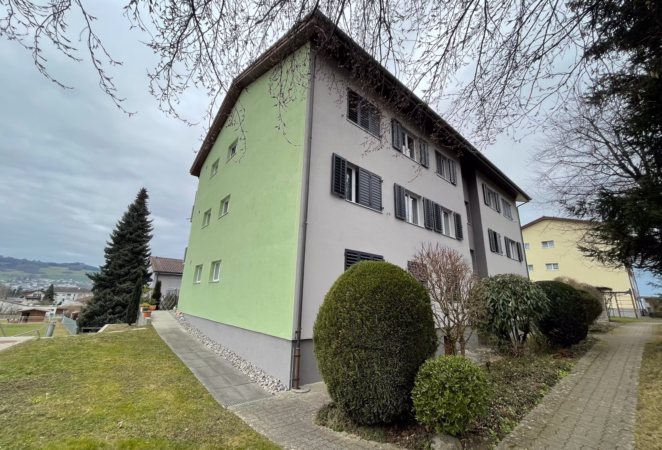 Seitenansicht eines gepflegten Mehrfamilienhauses mit grüner Fassade und Gartenanlage